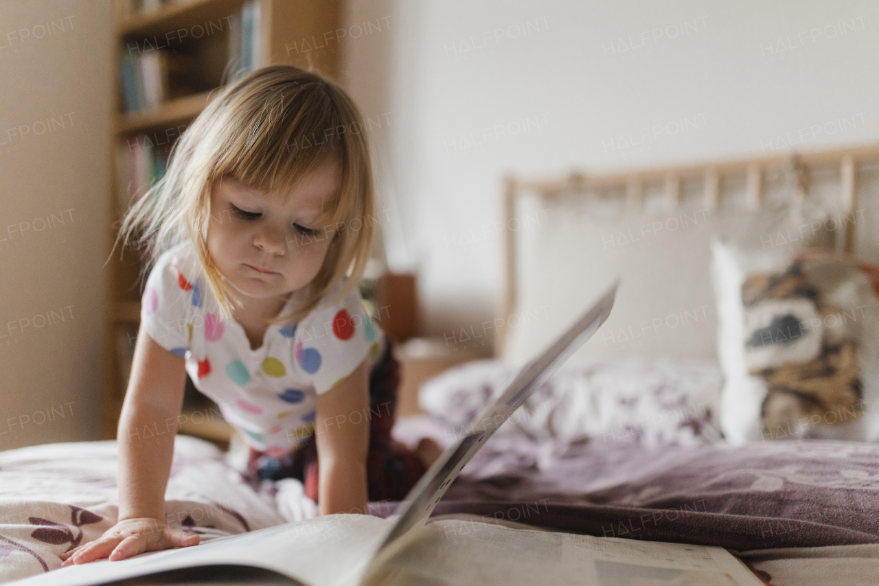 Cute little girl with bangs reading book lying on bed in her child's room.