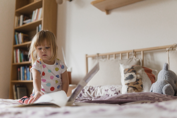 Cute little girl with bangs reading book lying on bed in her child's room.