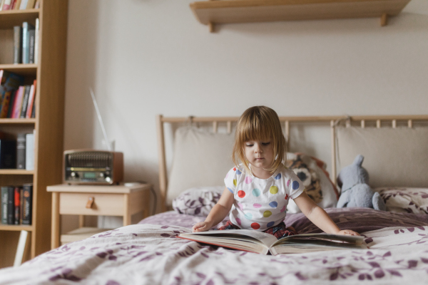 Cute little girl with bangs reading book lying on bed in her child's room.