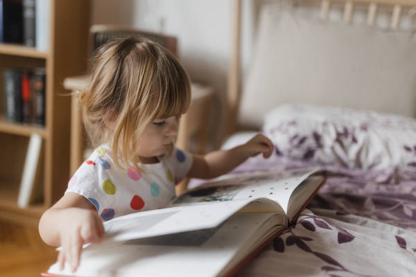 Cute little girl with bangs reading book lying on bed in her child's room.