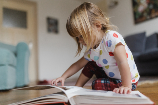 Cute little girl with bangs reading book sitting on the floor in living room.