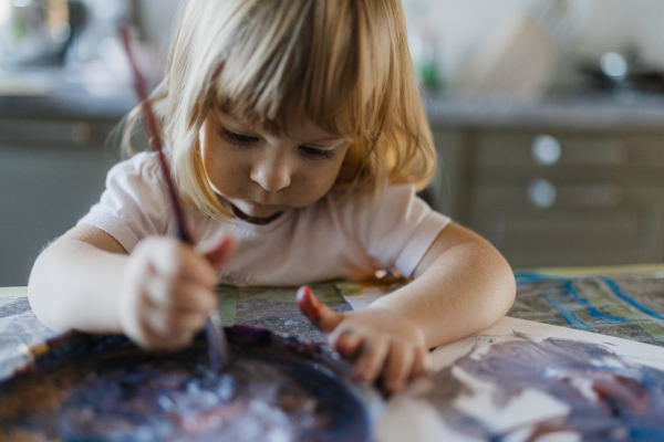 Cute little girl painting with tempera paint at home, using a paintbrush.