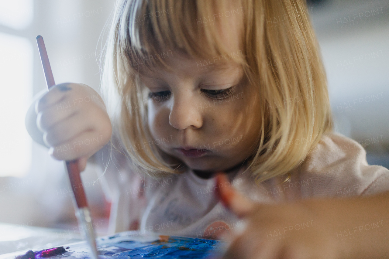 Cute little girl painting with tempera paint at home, using a paintbrush.