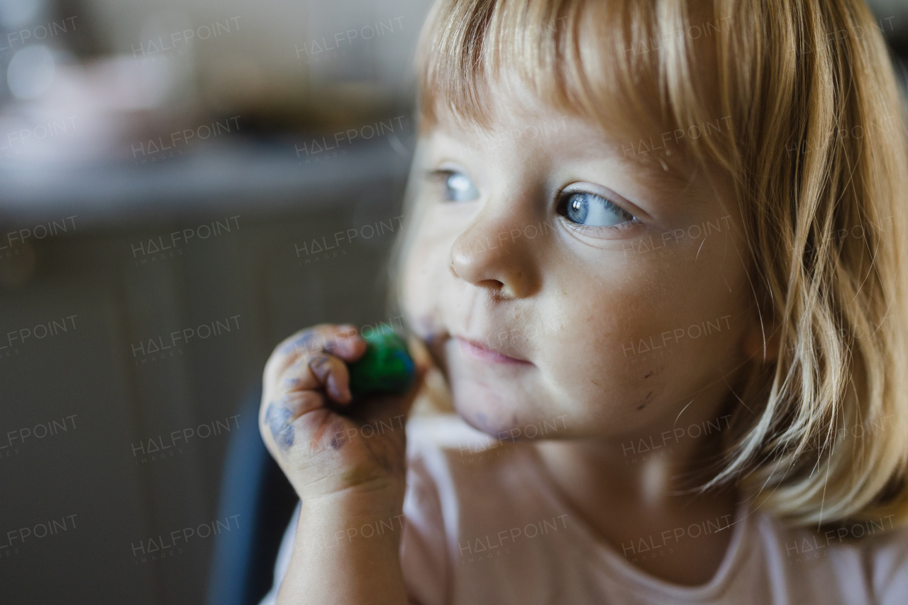 Portrait of cute little girl playing with playdough. Rolling, squashing playdough on cheek.