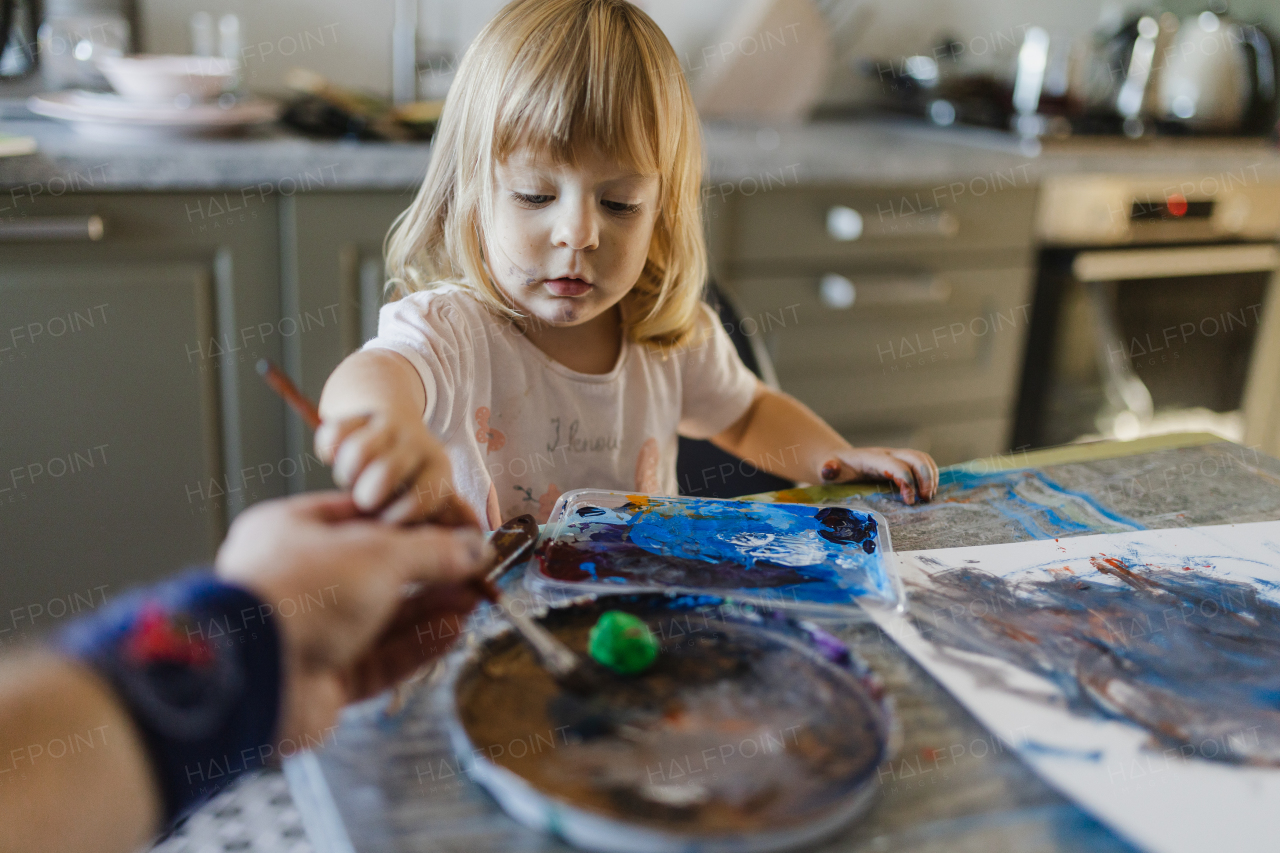 Father helping little girl painting with tempera paint at home, using a paintbrush.