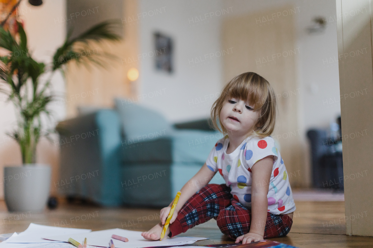 Cute little girl drawing with crayons, sitting on the floor in the living room. Outlining her own foot on paper.