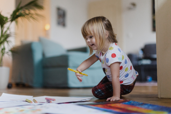 Cute little girl drawing with crayons, sitting on the floor in the living room.