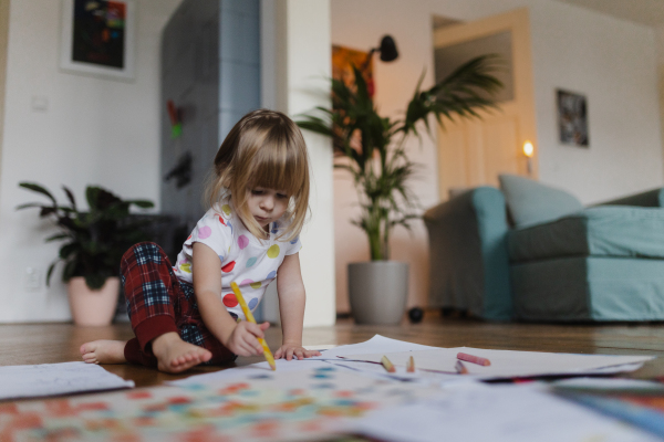 Cute little girl drawing with crayons, sitting on the floor in the living room.