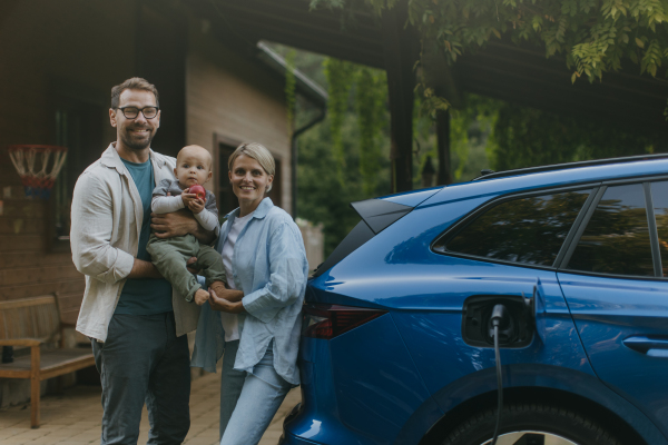 Young family with little baby standing by their electric car. Electric vehicle with charger in charging port.