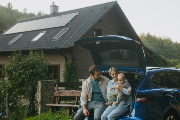 Family sitting in the trunk of electric car, with house with solar panels on roof behind them. Solar energy and sustainable lifestyle of young family. Concept of green energy and sustainable future for next generations.