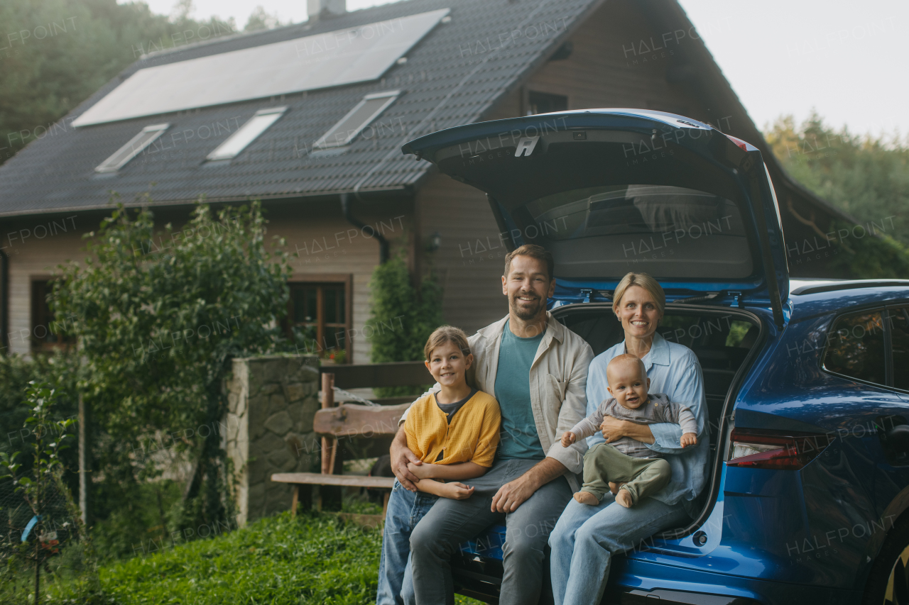 Family sitting in the trunk of electric car, with house with solar panels on roof behind them. Solar energy and sustainable lifestyle of young family. Concept of green energy and sustainable future for next generations.