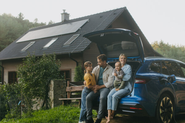 Family with electric car standing in front their house with solar panels on the roof. Solar energy and sustainable lifestyle of young family. Concept of green energy and sustainable future for next generations.