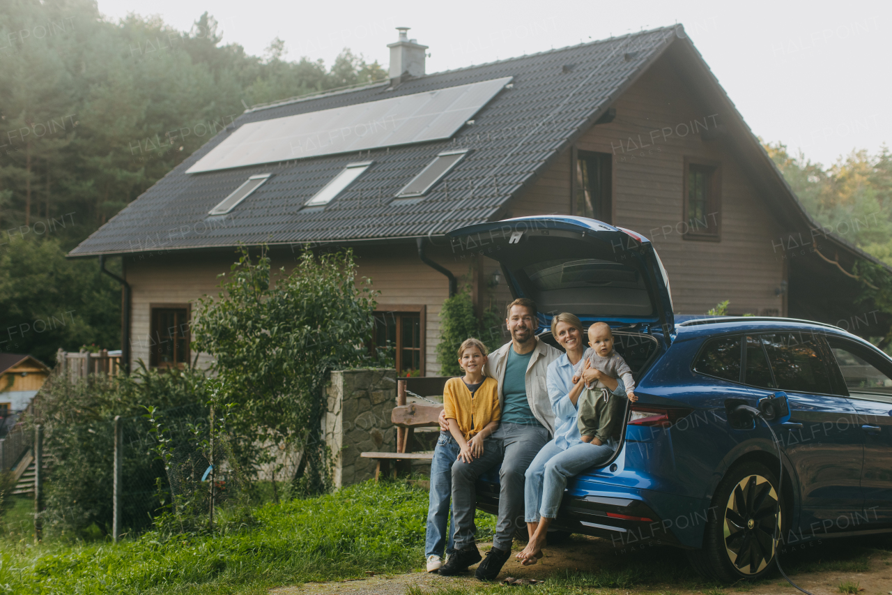 Family with electric car standing in front their house with solar panels on the roof. Solar energy and sustainable lifestyle of young family. Concept of green energy and sustainable future for next generations.