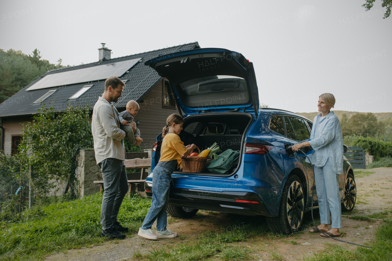 Family with electric car standing in front their house with solar panels on the roof. Solar energy and sustainable lifestyle of young family. Concept of green energy and sustainable future for next generations.