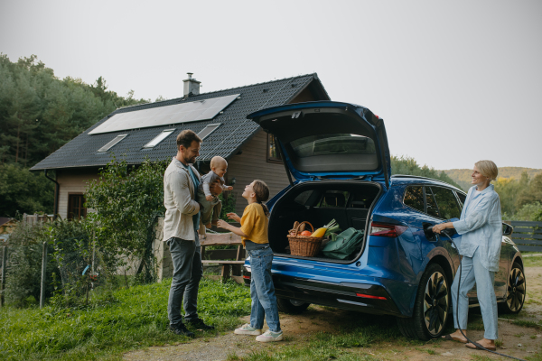 Family with electric car standing in front their house with solar panels on the roof. Solar energy and sustainable lifestyle of young family. Concept of green energy and sustainable future for next generations.