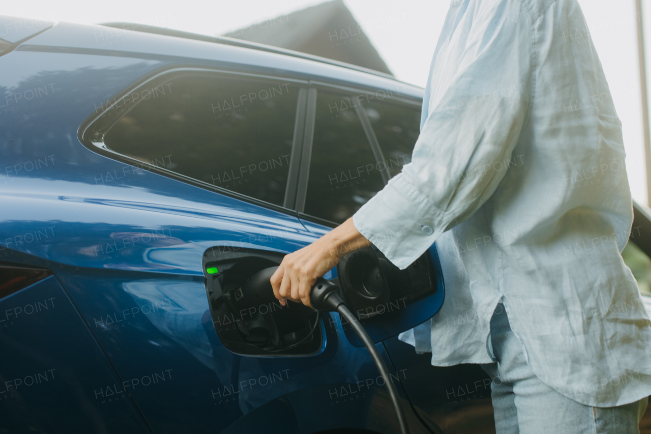 Portrait of a woman charging electric car in front of house, plugging the charger into the charging port, unplugging charger from the fully charged car before going to the office.