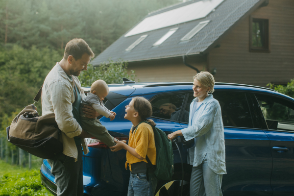 Family with electric car standing in front their house with solar panels on the roof. Solar energy and sustainable lifestyle of young family. Concept of green energy and sustainable future for next generations.