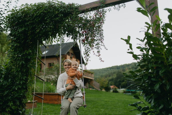 Beautiful mother and baby swinging on a swing in the garden, having fun and spending quality time together outdoors during a warm autumn day.