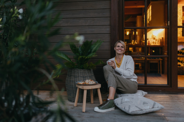 Woman sitting on the terrace during dusk, sipping warm tea and enjoying the view. Inside the house, it's warm and inviting, while she is dressed in a cozy sweater, sitting on soft pillows. Hygge atmosphere.