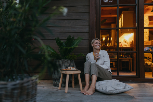 Woman sitting on the terrace during autumn dusk, sipping warm tea and enjoying the view. Inside the house behind her, it's warm and inviting, while she is dressed in a cozy sweater, sitting on soft pillows. Autumn hygge atmosphere.