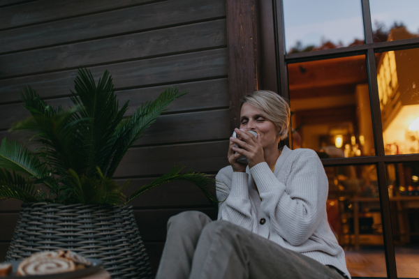 Low angle of woman sitting on the terrace during autumn dusk, sipping warm tea and enjoying the view. Inside the house behind her, it's warm and inviting, while she is dressed in a cozy sweater, sitting on soft pillows. Autumn hygge atmosphere.