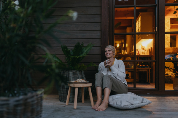 Woman sitting on the terrace during dusk, sipping warm tea and enjoying the view. Inside the house, it's warm and inviting, while she is dressed in a cozy sweater, sitting on soft pillows. Hygge atmosphere.