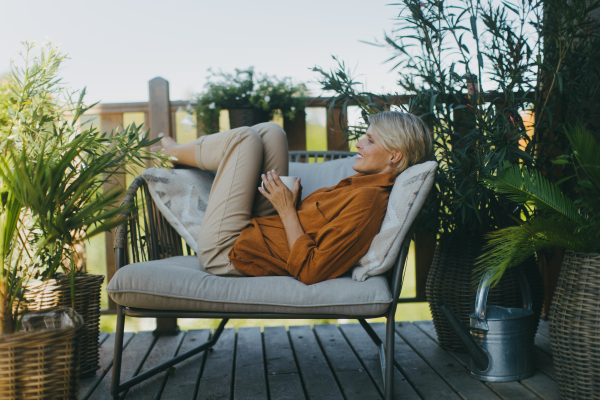 Beautiful woman relaxing in garden, sitting on patio chair and drinking cup of coffee. Mother having a moment to herself while her child is sleeping.