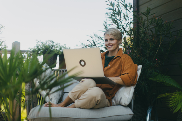 Woman in garden working on laptop, sitting on patio chair. Businesswoman working remotely from outdoor home office, thinking about new business or creative idea. Concept of green outdoor home office.