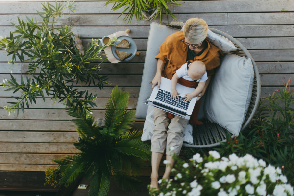 Top view of mother holding baby while working on laptop in garden. Businesswoman working remotely from outdoor home office and taking care of little son. Life work balance with kid. Working mom with toddler.