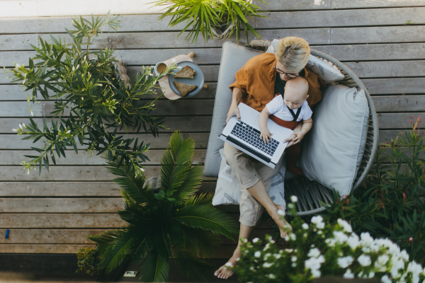 Top view of mother holding baby while working on laptop in garden. Businesswoman working remotely from outdoor home office and taking care of little son. Life work balance with kid. Working mom with toddler.