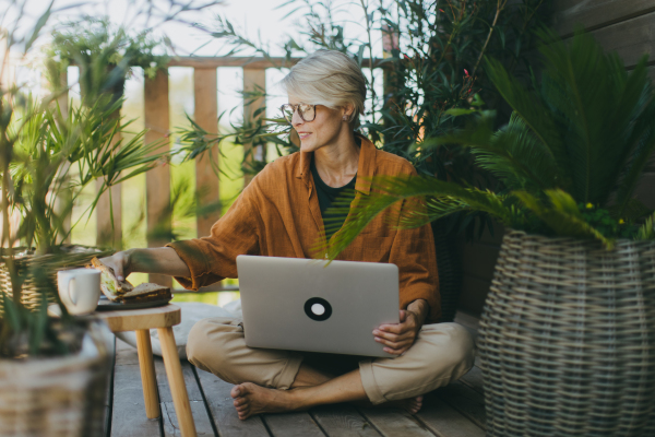 Woman working in garden, with laptop on legs. Businesswoman working remotely from the outdoor homeoffice, eating light lunch, sandwich. Concept of outdoor home office.