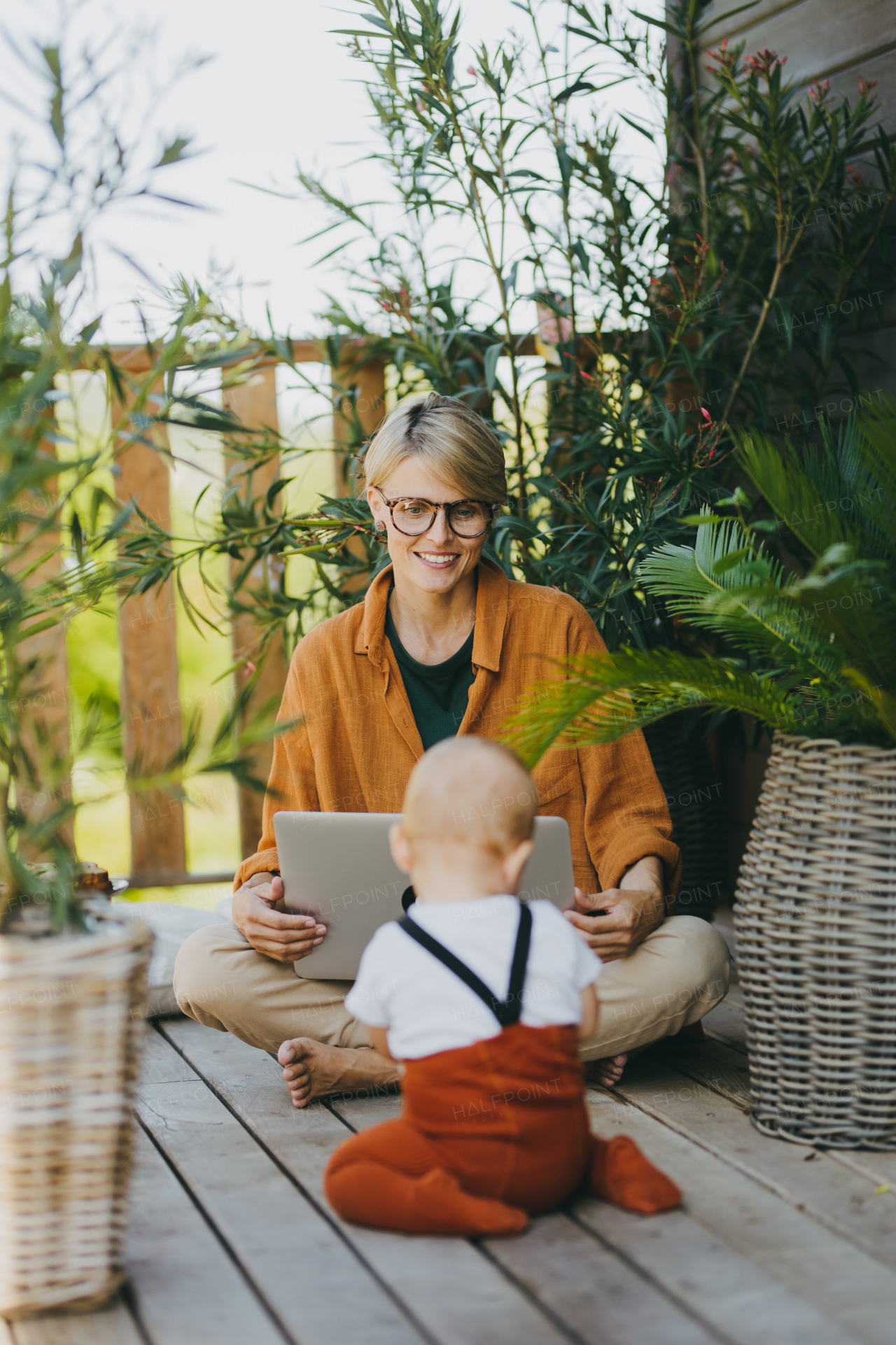 Mother holding baby while working on laptop in garden. Businesswoman working remotely from outdoor home office and taking care of little son. Life work balance with kid. Working mom with toddler.