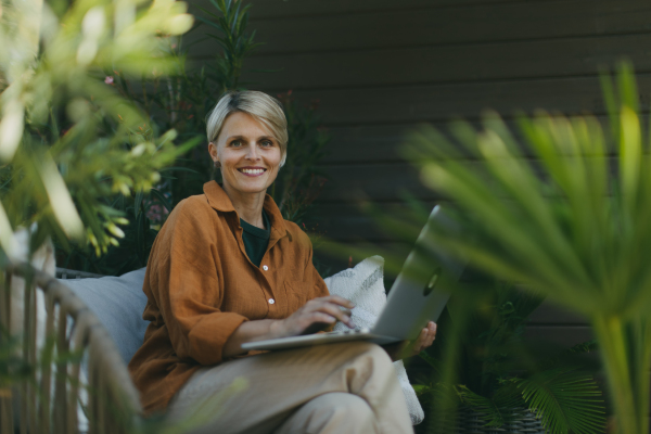 Woman in garden working on laptop, sitting on patio chair. Businesswoman working remotely from outdoor home office, thinking about new business or creative idea. Concept of green outdoor home office.