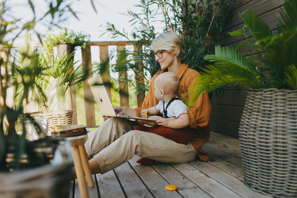 Mother holding baby while working on laptop in garden. Businesswoman working remotely from outdoor home office and taking care of little son. Life work balance with kid. Working mom with toddler.