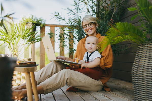 Mother holding baby while working on laptop in garden. Businesswoman working remotely from outdoor home office and taking care of little son. Life work balance with kid. Working mom with toddler.