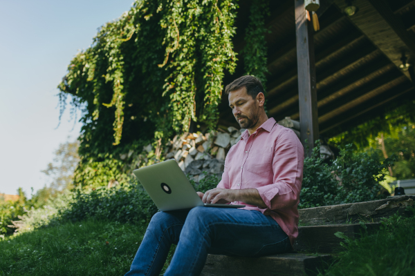 Low angle shot of man working outdoors in the garden, with laptop on his legs. Businessman working remotely from homeoffice, thinking about new business or creative idea.