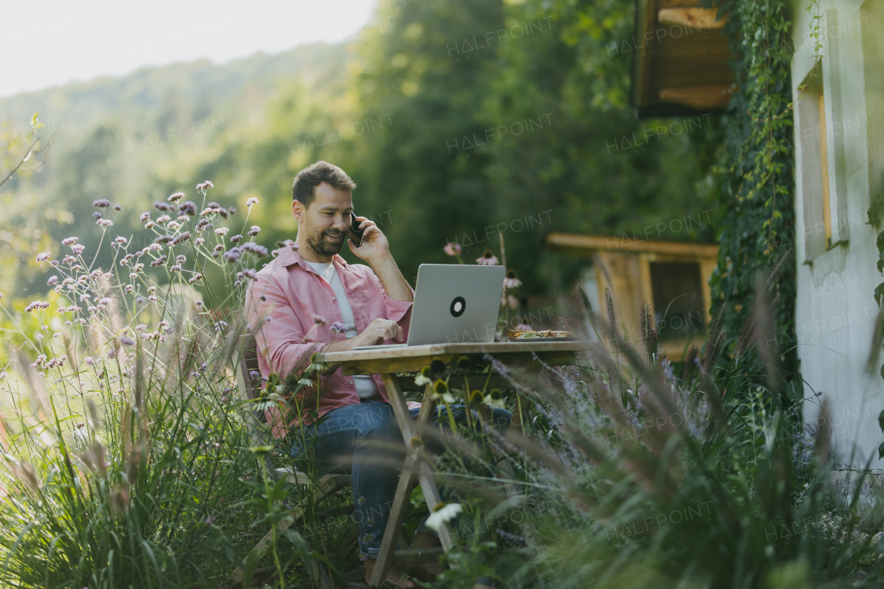 Man working in the garden on laptop, phone calling on smartphone. Businessman working remotely from outdoor homeoffice, thinking about new business or creative idea.