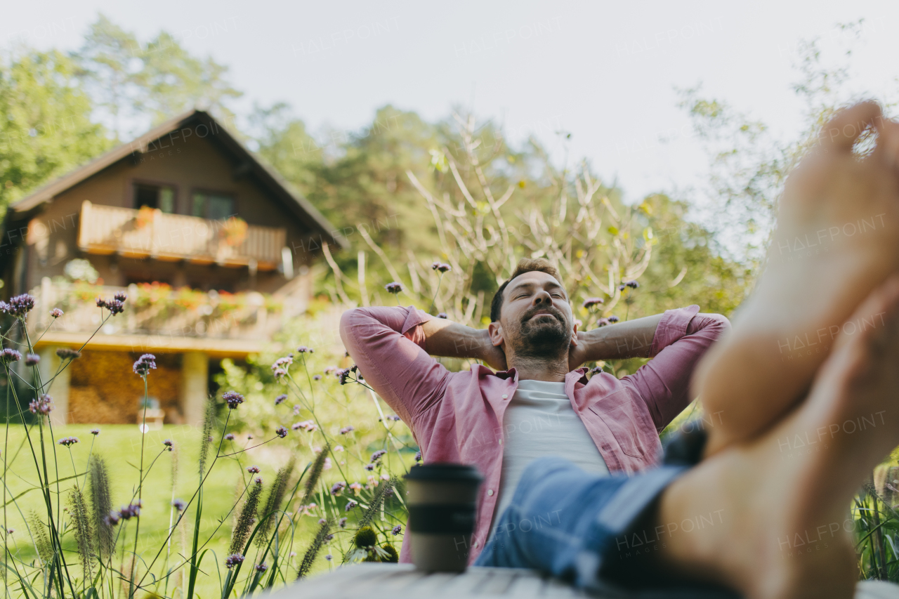 Relaxed barefoot man sitting in the garden with feet up on the table. Father having moment to himself while his kids are in school.