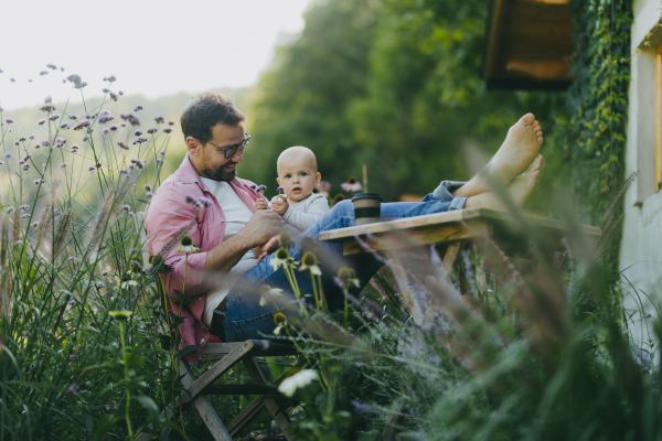 Man sitting in the garden with feet on table, playing with his baby son. Father having bonding moment with his little cute kid. Baby playing with flower.