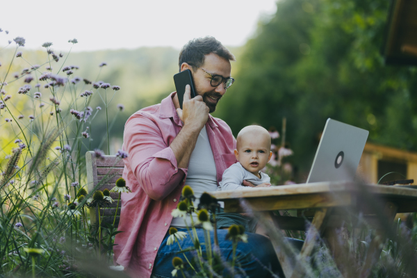 Father holding baby while working on laptop outdoors, in the garden. Businessman working remotely from homeoffice, making phone call with client and taking care of little son. Life work balance with kid.