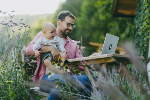 Father holding baby while working on laptop in the garden. Businessman working remotely from outdoor homeoffice and taking care of little son. Life work balance with kid.