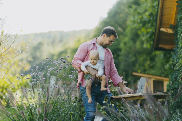 Father holding baby while working on laptop in the garden. Businessman working remotely from outdoor homeoffice and taking care of little son. Life work balance with kid.