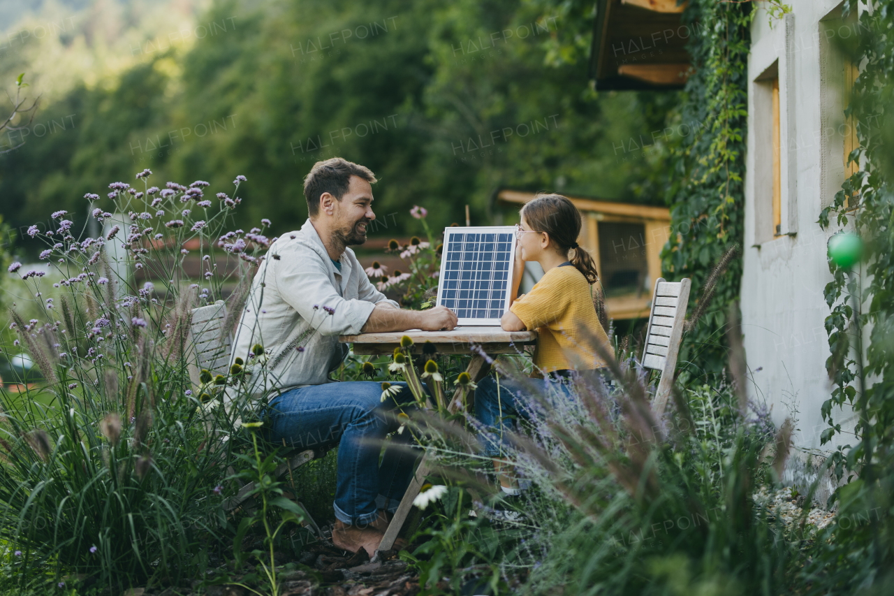 Father with daughter sitting outdoors in the garden, with model of solar panel. Solar energy and sustainable lifestyle of young family. Concept of green energy and sustainable future for next generations.