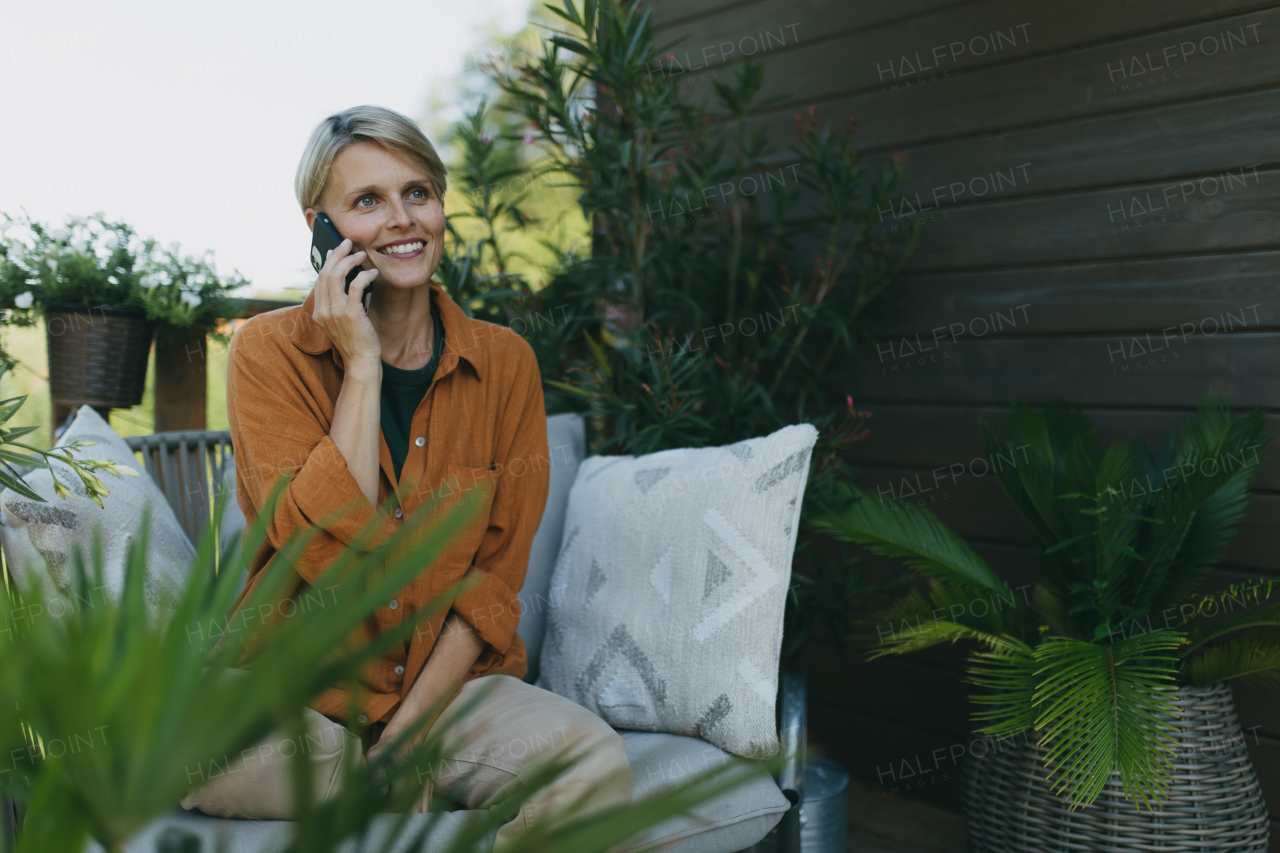 Beautiful woman making call with smart phone outdoors, sitting on patio chair. Mother having moment to herself while her child is sleeping.