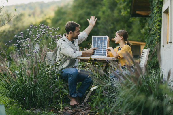 Father with daughter sitting outdoors in the garden, with model of solar panel. Solar energy and sustainable lifestyle of young family. Concept of green energy and sustainable future for next generations.