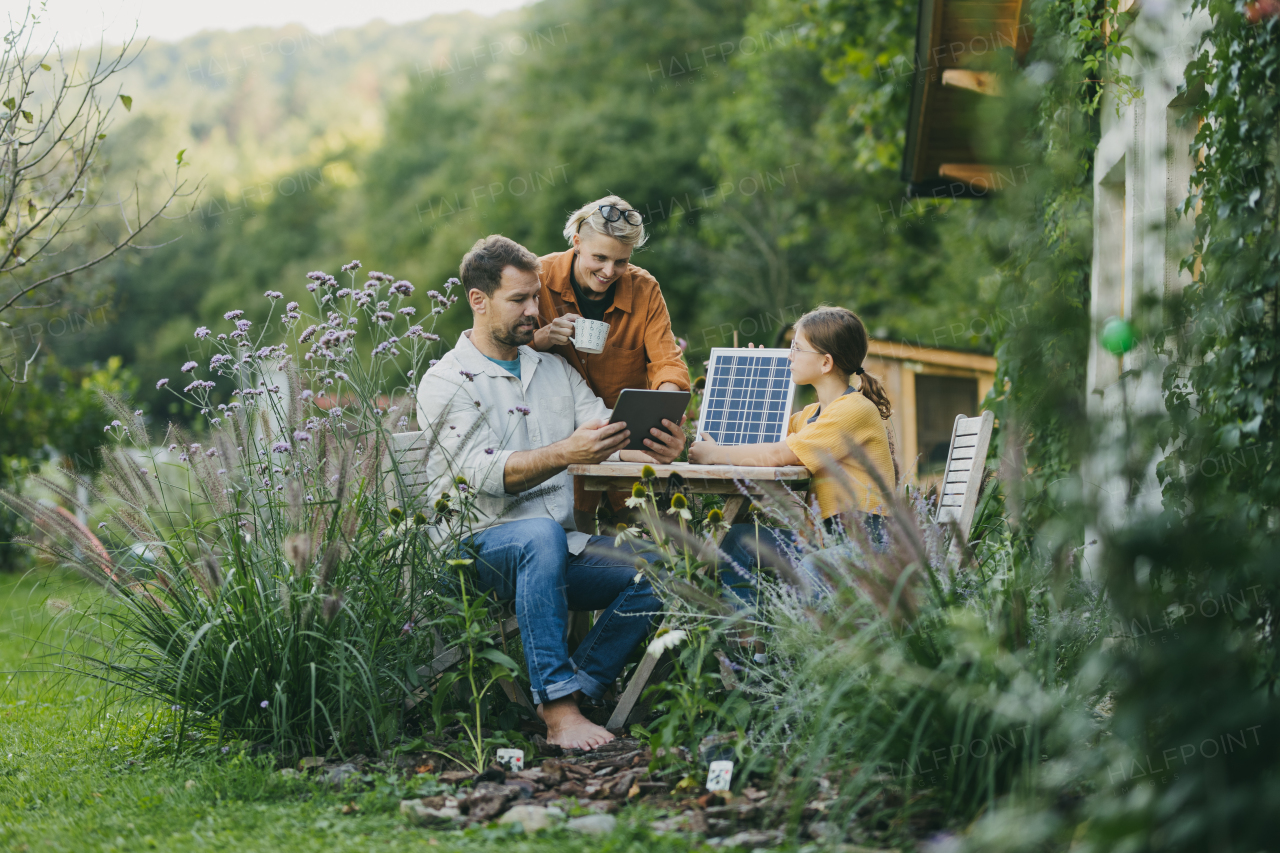 Parents with daughter sitting outdoors in the garden, with model of solar panel, learning about solar energy. Solar energy and sustainable lifestyle of young family. Concept of green energy and sustainable future for next generations.