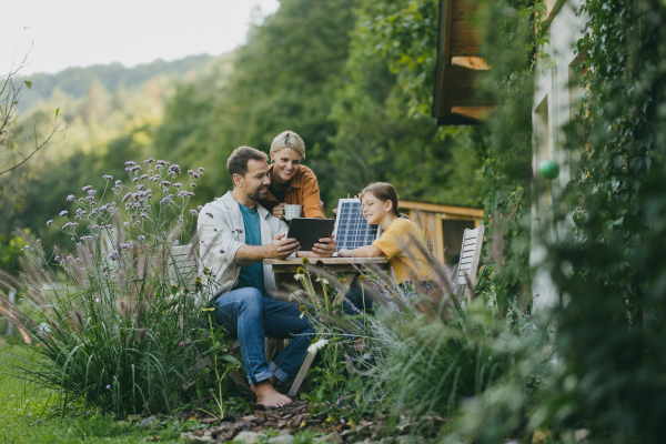 Parents with daughter sitting outdoors in the garden, with model of solar panel, learning about solar energy. Solar energy and sustainable lifestyle of young family. Concept of green energy and sustainable future for next generations.