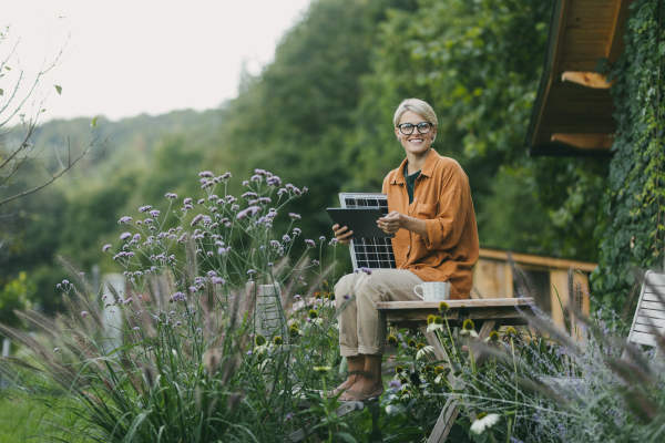 Woman in garden holding solar panel and tablet. Looking for solar panels grants, funds for homeowners. Solar energy and sustainable lifestyle for the family in house.