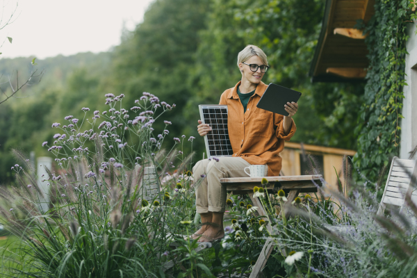 Woman in garden holding solar panel and tablet. Looking for solar panels grants, funds for homeowners. Solar energy and sustainable lifestyle for the family in house.