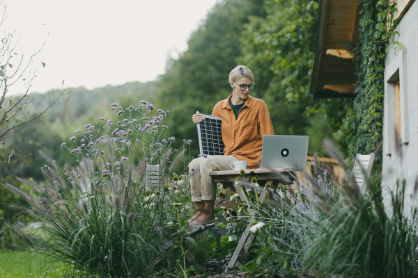 Woman in garden holding solar panel. Looking for solar panels grants, funds for homeowners on laptop. Solar energy and sustainable lifestyle for the family in house.